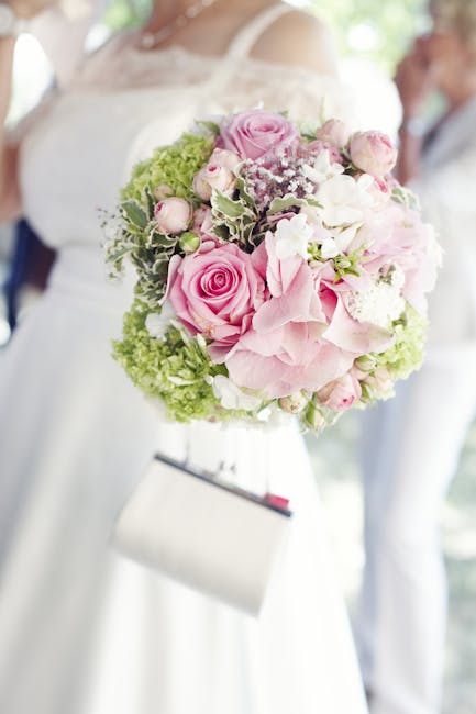 A bride in a white dress holding a round bouquet featuring soft pink roses, white hydrangeas, and delicate baby’s breath, accented with green foliage. The bouquet is wrapped in light pink paper and 