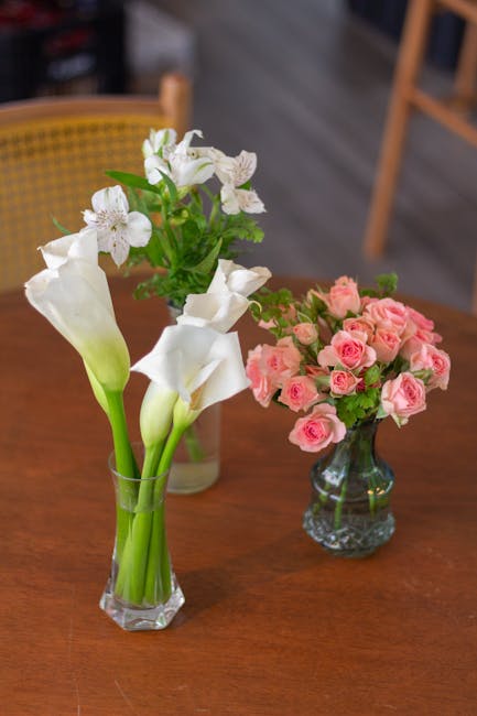 A clear glass vase containing a bouquet of white calla lilies with smooth, curved petals and elongated stems, alongside a separate small glass vase filled with a lush arrangement of pink roses in full