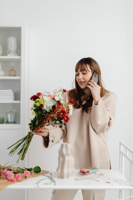 A woman with long brown hair and bangs, dressed in a cream-colored sweater, is standing at a white table in a bright, minimalistic interior. She is holding a large, vibrant bouquet of mixed fresh flow