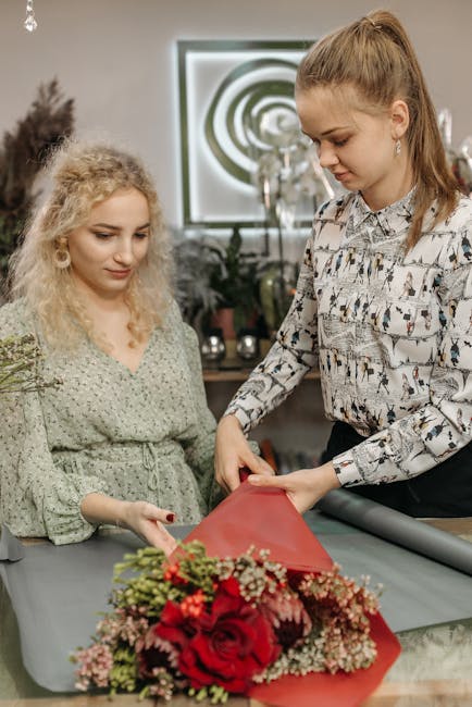 Two women are preparing a floral gift indoors, with one woman holding a bouquet of fresh roses, eustoma, and baby's breath wrapped in red and pink wrapping paper on a table. The woman on the left has 