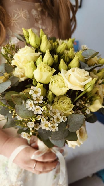 A close-up of a bridal bouquet featuring soft white and pale yellow roses, complemented by small white waxflowers and sprigs of eucalyptus leaves. The bouquet is held by a person wearing a wedding dre