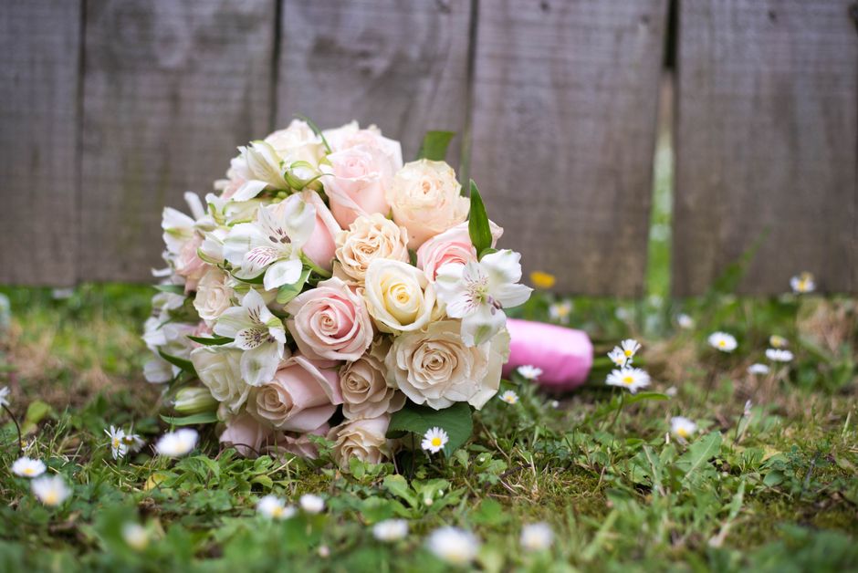 A bouquet of fresh pale pink and cream roses, accented with white lilies and green foliage, resting on grass with small white daisies scattered around. The bouquet is wrapped in soft pink paper and ti