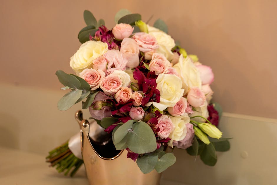 A floral arrangement featuring a bouquet of fresh flowers including white and pale pink roses, with some darker pink or burgundy blooms and green eucalyptus leaves, arranged in a rounded style. The fl