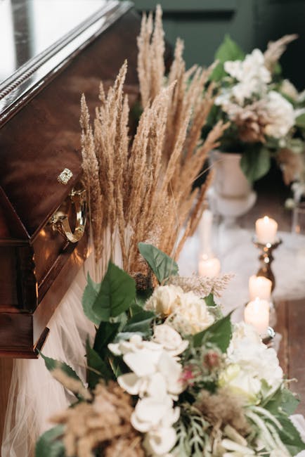 A floral arrangement placed on a wooden surface beside a dark polished musical instrument, featuring a bouquet of white and pale pink roses, white orchids, and green foliage. Behind the bouquet, tall,