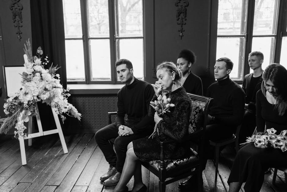 A black-and-white photograph showing a group of six people seated indoors during a funeral service. The setting features large windows with natural light illuminating the room, dark wooden floors, and