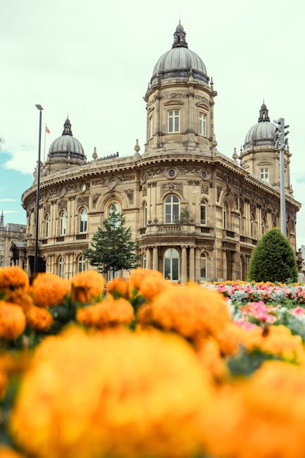 A close-up view of vibrant orange marigold flowers in the foreground, with a blurred background showing an ornate historic building featuring classical architecture, large arched windows, decorative s