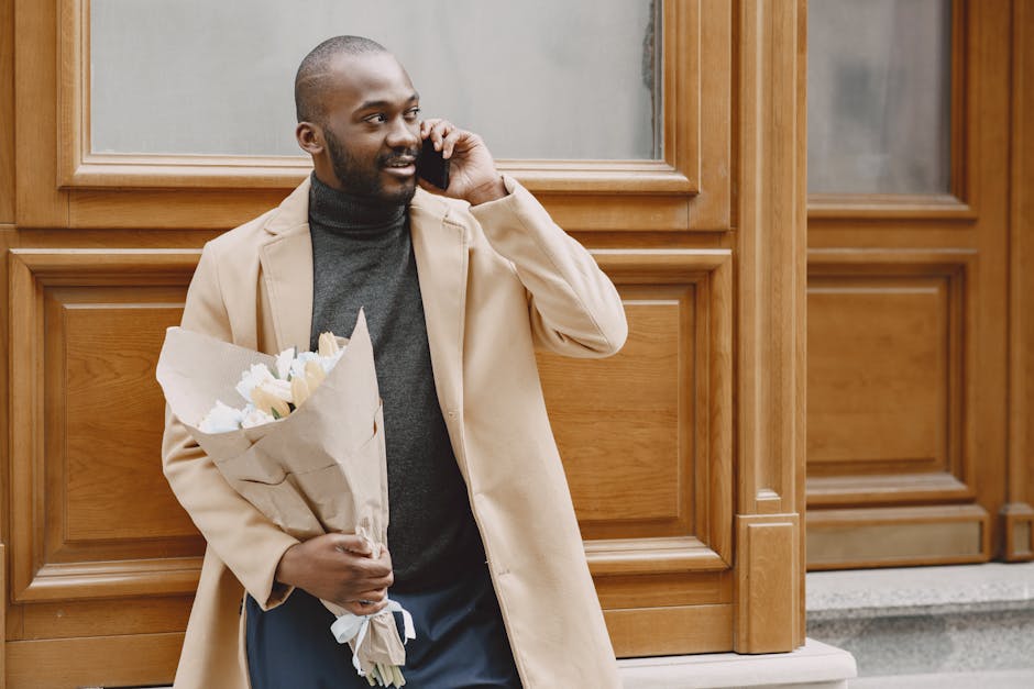 A man with dark skin and a beard is standing outdoors in front of a wooden paneled wall, holding a bouquet of white tulips wrapped in brown paper. He is wearing a beige overcoat over a dark gray turtl