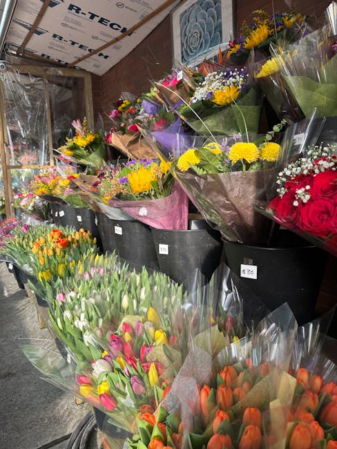 A variety of fresh flower bouquets arranged in black buckets and wrapped in colored paper and cellophane, displayed inside a flower shop. The arrangements include bright yellow chrysanthemums, multico
