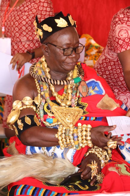 The image features an elderly woman dressed in traditional African attire, adorned with elaborate gold jewelry including necklaces, bracelets, and a headpiece, sitting indoors on a red and patterned f